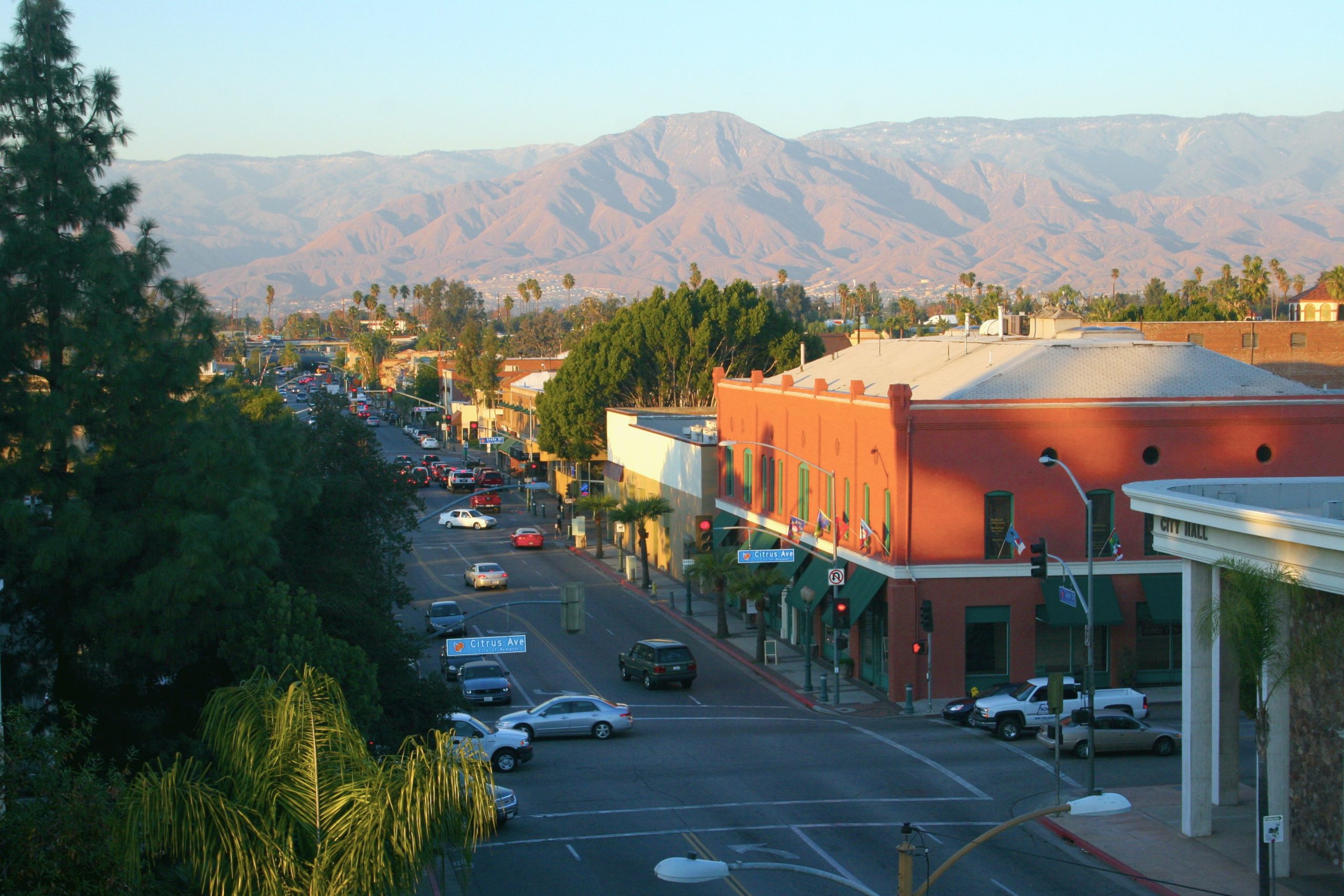 Aerial view of a bustling street lined with colorful buildings and palm trees. In the background, majestic mountains are lit by warm sunlight under a clear sky.