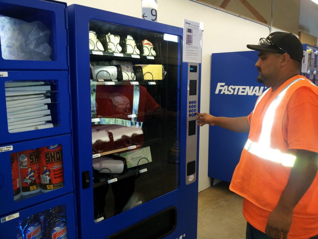 A man in a safety vest uses a blue vending machine stocked with safety gear and supplies in an indoor setting.