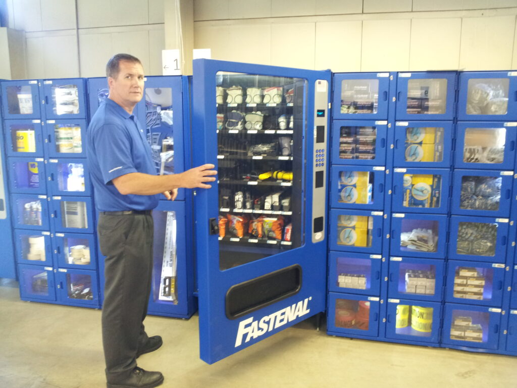 A man in a blue shirt opens a Fastenal vending machine filled with industrial supplies, with additional supply lockers in the background.