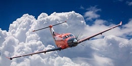 A bright red airplane soars through a clear blue sky, surrounded by large, fluffy white clouds, conveying a sense of adventure and freedom.