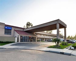 Single-story hotel with brown columns and a red roofed entrance, set against a bright sky. The surroundings include neatly trimmed bushes and parked cars.