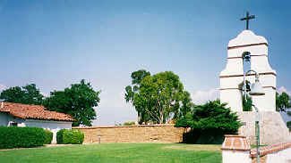 A serene landscape with a white bell tower topped by a cross, surrounded by lush greenery and a small building with a red-tiled roof under a clear blue sky.
