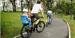 A man cycles with a child in a blue seat on a park path, followed by another cyclist. Trees and grass surround the path, conveying a serene, leisurely mood.