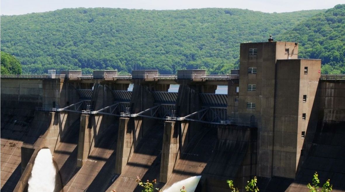 A large concrete dam with multiple spillways releases water, framed by lush green trees and hills. The scene conveys a sense of engineering amidst nature.