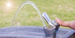 Close-up of a hand pressing a silver water fountain with water arcing upward. The background shows a grassy field lit by soft sunlight.