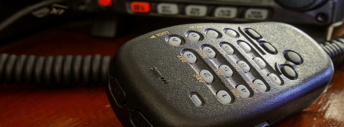 Close-up of a black handheld radio microphone with buttons on a wood surface. Background shows part of the radio unit with control buttons.
