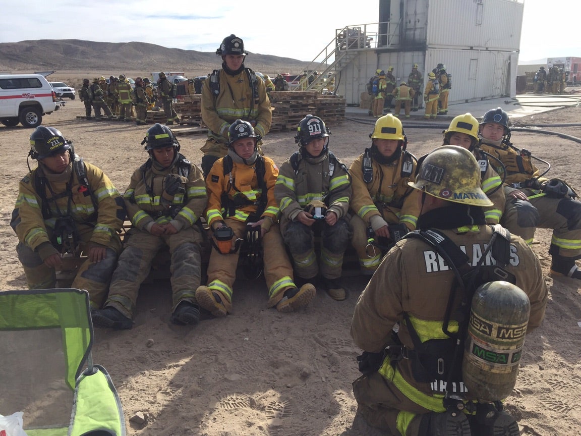 A group of firefighters in gear sitting on the ground in a desert setting, appearing tired but engaged. More firefighters and equipment are visible in the background.