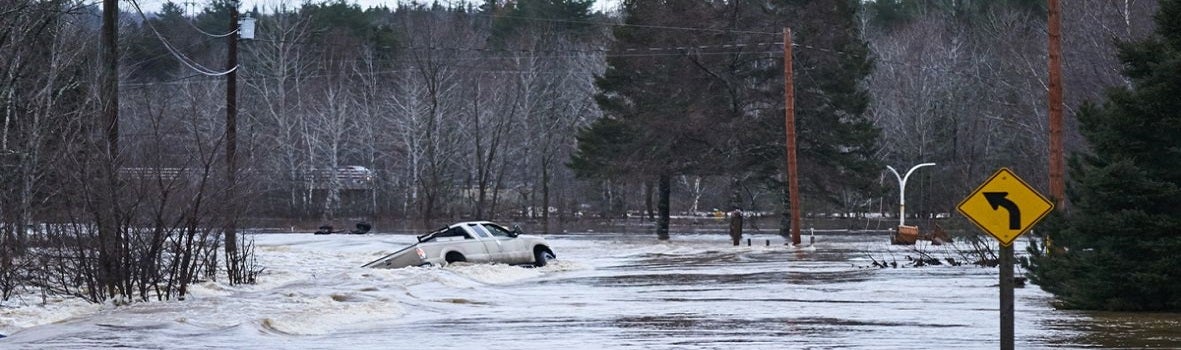 A flooded road with a partially submerged white truck near a curve sign. Surrounding trees and power poles stand in murky water under a gray sky.