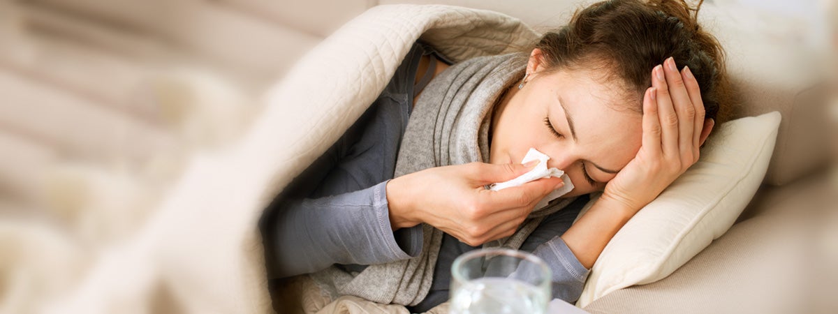 A woman lies in bed, appearing sick and exhausted. She holds a tissue to her nose, wrapped in a cozy blanket, with water on a bedside table.
