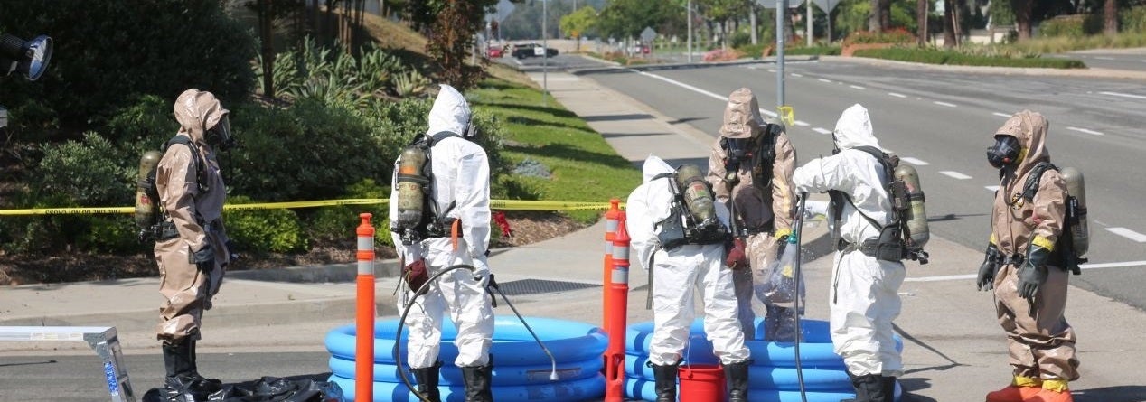 Seven people in hazmat suits work on a closed-off street with blue decontamination pools. The scene conveys urgency and caution in a potential hazard situation.