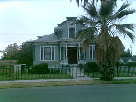 A quaint, two-story gray Victorian house with white trim, a prominent palm tree in front, and a black iron fence. The setting appears calm and residential.