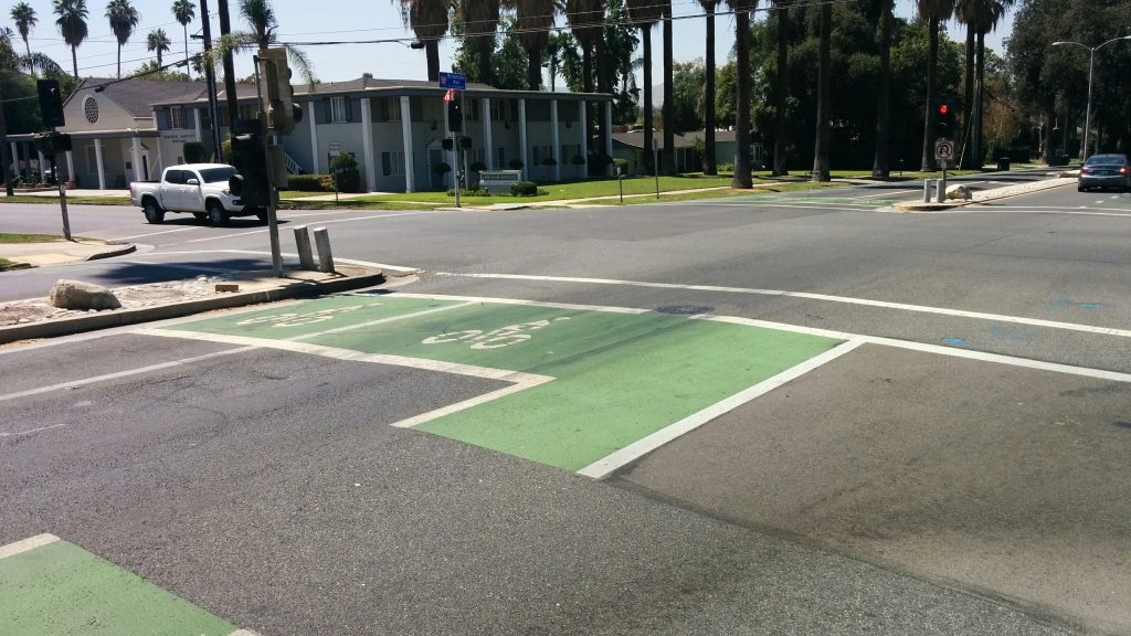 A sunny street intersection with marked bike lanes in bright green. Palm trees and residential buildings are visible; a white truck waits at the light.