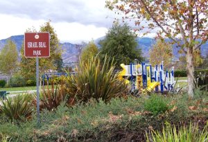 Playground at Israel Beal Park with colorful slides and climbing structures, surrounded by lush greenery and mountains under a cloudy sky.