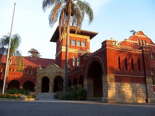 Historic red brick building with arches and a tower, surrounded by palm trees. The evening light casts a warm glow, creating a serene atmosphere.