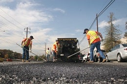 Workers in orange vests pave a road using a large machine. They spread asphalt under a clear sky, creating a sense of teamwork and progress.