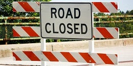 "Road closed sign with bold black letters on a white background, framed by orange and white striped barricades. Trees visible in the background."