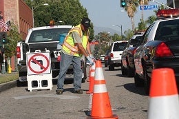 Worker in a safety vest places orange traffic cones on a road. A no-turn sign is visible, and cars are lined up. The scene conveys road maintenance work.