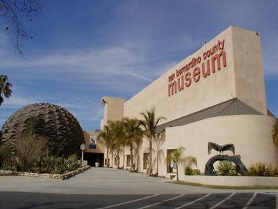 A large beige building labeled "San Bernardino County Museum," with a geodesic dome, palm trees, and blue sky. The setting is calm and inviting.
