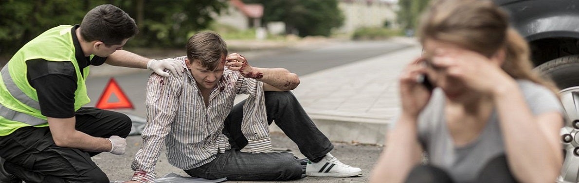 A distressed man sits injured on the roadside, attended by an EMT in a reflective vest. Nearby, a woman makes a phone call, likely seeking help.