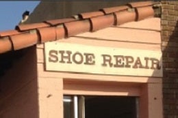 A quaint sign reading "Shoe Repair" hangs under a roof with rustic red tiles. The setting appears sunny, evoking a vintage, small-town charm.