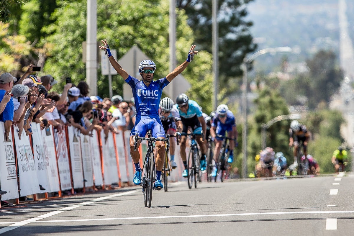 Cyclist in blue, arms raised in victory, crosses the finish line first. A cheering crowd lines the road, with more cyclists trailing behind.