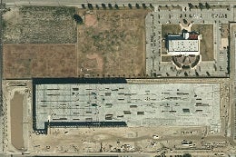 Aerial view showing a rectangular industrial building under construction with a completed roof, adjacent to a partially paved lot and surrounding fields.