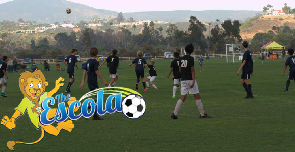 A youth soccer match is taking place on a grassy field with hills in the background. A cartoon lion and “The Escola” logo with a soccer ball are in the foreground.