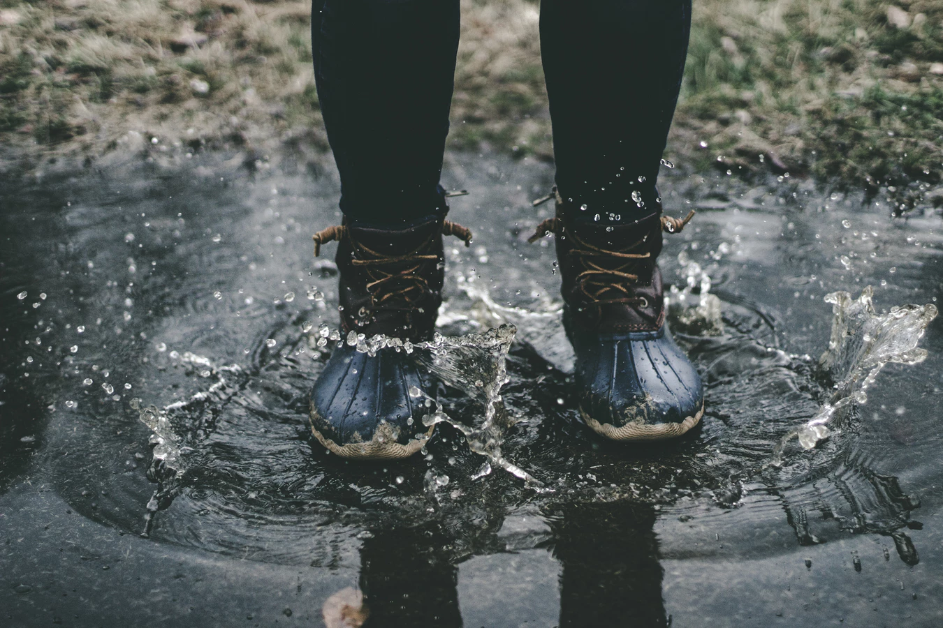 A person wearing dark boots jumps in a puddle, causing water to splash energetically. The scene conveys a playful, carefree mood on a damp day.
