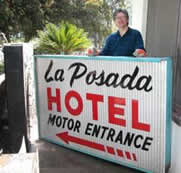 A person stands smiling beside a vintage sign reading "La Posada Hotel Motor Entrance" with red and black text. Sunlit greenery surrounds the scene.