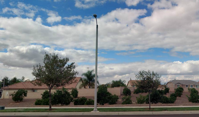 A street with a single lamppost stands between two small trees under a partly cloudy sky. Houses and a stone wall line the background, conveying a suburban feel.