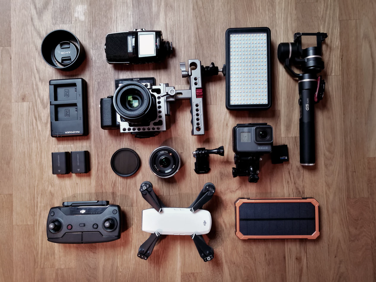 Camera gear flat lay on wooden floor: camera lenses, professional camera, drone, battery packs, gimbal, LED light, and remote controller.