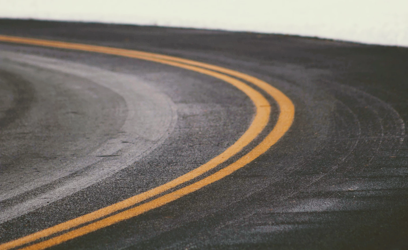 Curved road with double yellow lines, highlighting the road's arc. The asphalt is dark with visible tire marks, creating a mood of movement and direction.