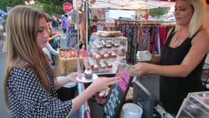 A woman buys a cupcake from a vendor at an outdoor market stall. The stall displays cupcakes and colorful clothes. The scene is lively and inviting.