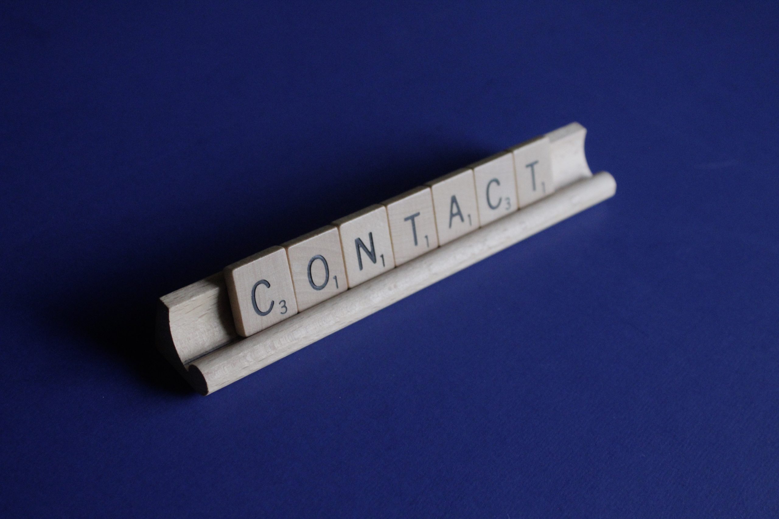 Wooden Scrabble tiles on a rack spell "CONTACT" against a deep blue background, conveying communication and connection.