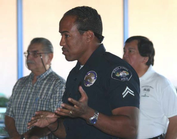 A police officer in uniform speaks, using hand gestures, alongside two men in casual attire. The scene appears to be a community meeting, conveying a serious tone.