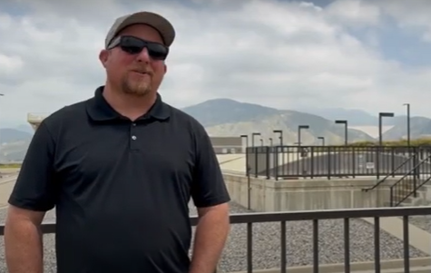 Man in sunglasses and a cap stands outdoors in front of a metal railing. Mountains and a cloudy sky form the backdrop, creating a calm atmosphere.