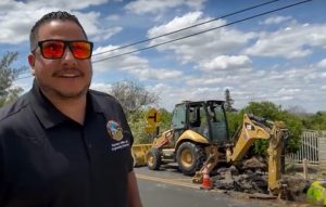 A smiling man in sunglasses stands on a sunny road beside construction, with a backhoe actively digging. Orange cones mark the area. Sky is partly cloudy.