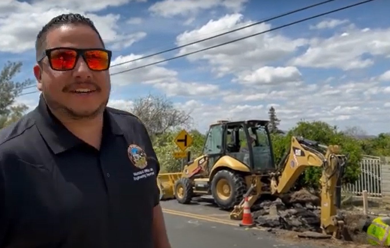 A smiling man in sunglasses stands on a sunny road beside construction, with a backhoe actively digging. Orange cones mark the area. Sky is partly cloudy.