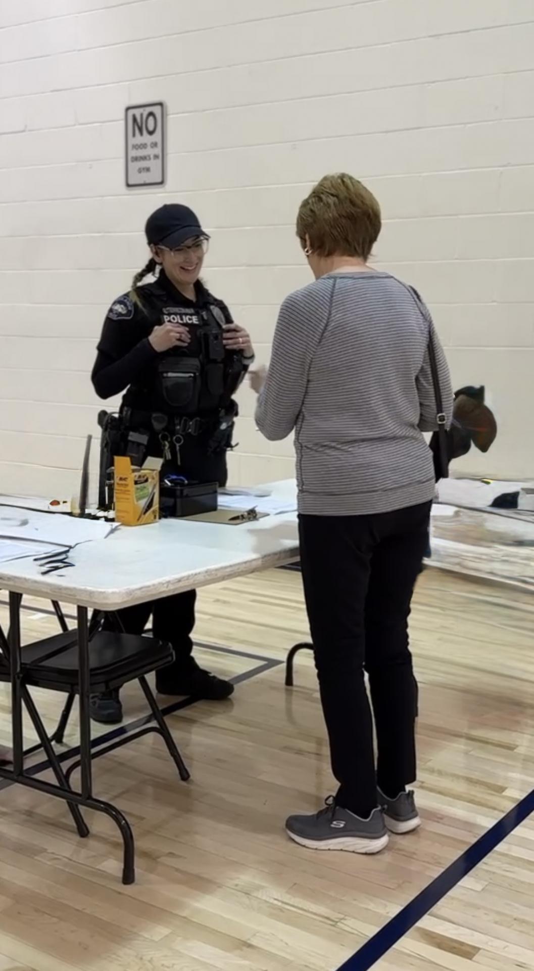 A police officer smiles while talking to a woman at a table indoors. Papers and folders are spread on the table. The setting appears friendly and informal.