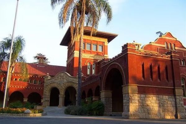 A historic red-brick building with arches, a tall central tower, and palm trees in front, photographed in daylight.