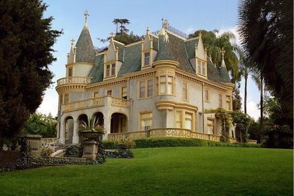 Large Victorian-style mansion with multiple gables, turrets, and ornate details, surrounded by greenery and palm trees on a well-manicured lawn.