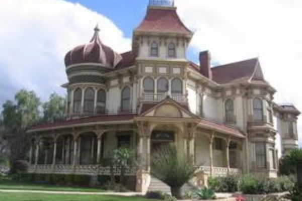 Large Victorian-style house with ornate trim, a wraparound porch, and a prominent round turret, set on a landscaped lawn under a partly cloudy sky.