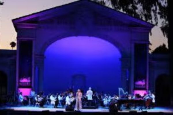 A symphony orchestra performs on an outdoor stage at dusk, illuminated by purple and blue lighting, with a singer standing at the front.