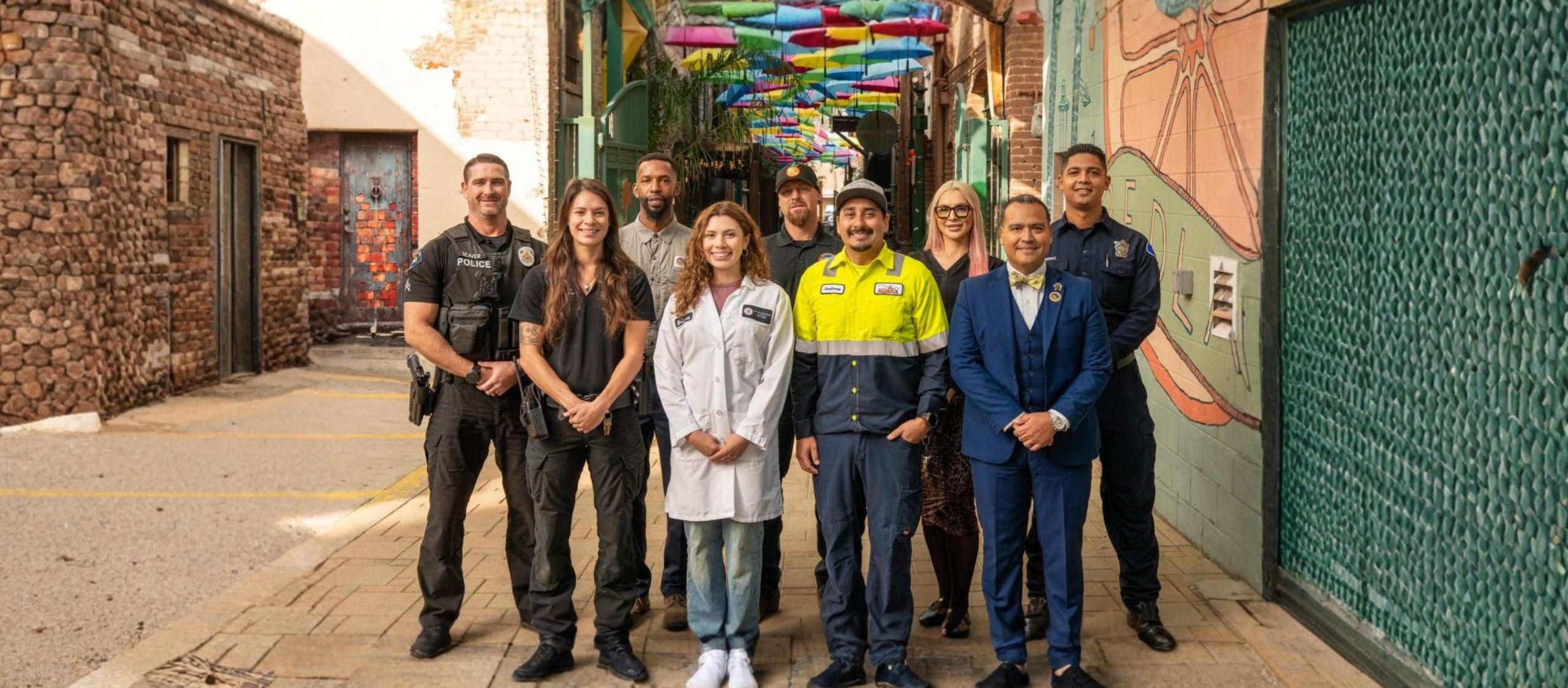 A group of nine people in various uniforms, including police, healthcare, and trade, stand in an outdoor alley with colorful umbrellas overhead.