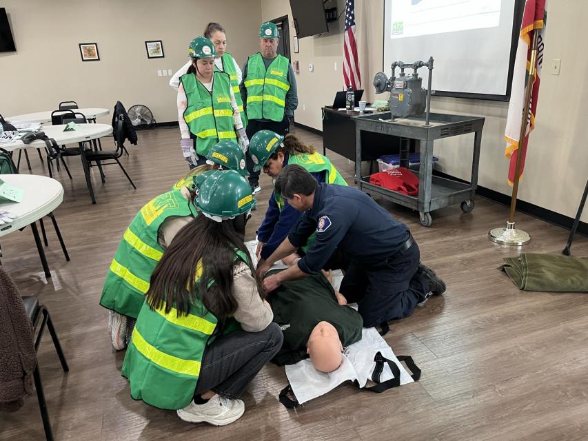 A group of people in green safety vests and helmets observe as an emergency responder demonstrates first aid on a training mannequin in a classroom setting.