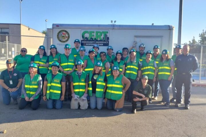 A group of people wearing green vests and hats pose in front of a CERT trailer outdoors during daylight.