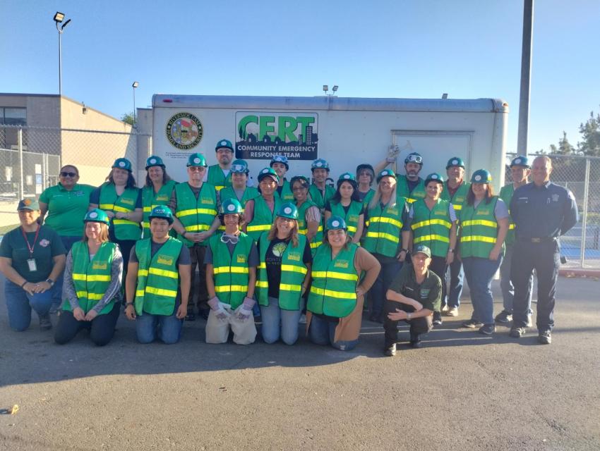 A group of people wearing green vests and hats pose in front of a CERT trailer outdoors during daylight.