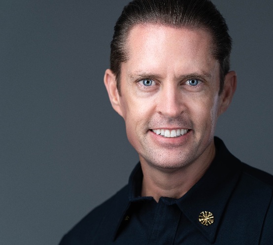 Man with short brown hair wearing a dark uniform shirt, smiling at the camera against a plain gray background.