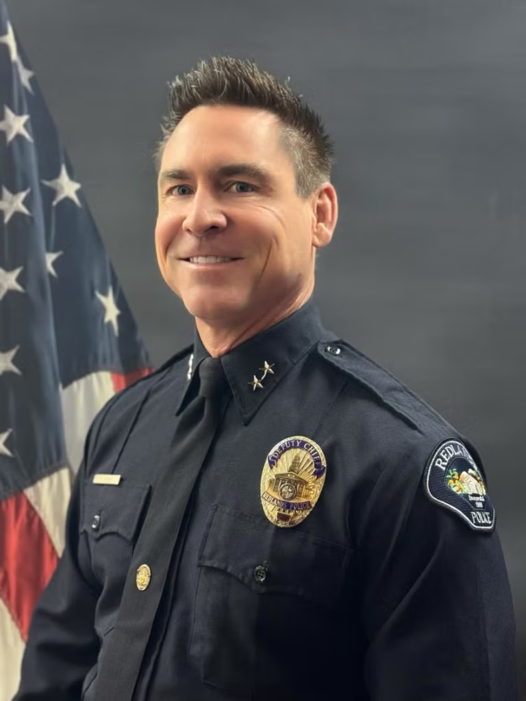 A police officer in uniform stands in front of an American flag, smiling at the camera. The badge and patch indicate affiliation with the Redlands Police Department.