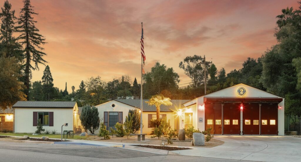 Redlands fire station no. 262 building with a flagpole in front, surrounded by trees, at sunset. The garage doors on the right are closed, with red lights visible inside.